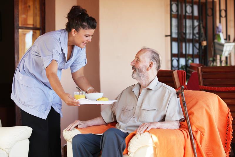 Carer in light blue uniform serving a meal to an elderly man in an armchair in a care home