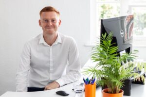 Sam Hickman sitting at a desk with a plant in the foreground.