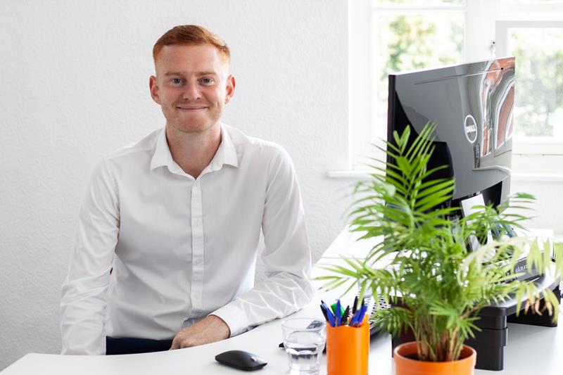 Sam Hickman sitting at a desk with a plant in the foreground.
